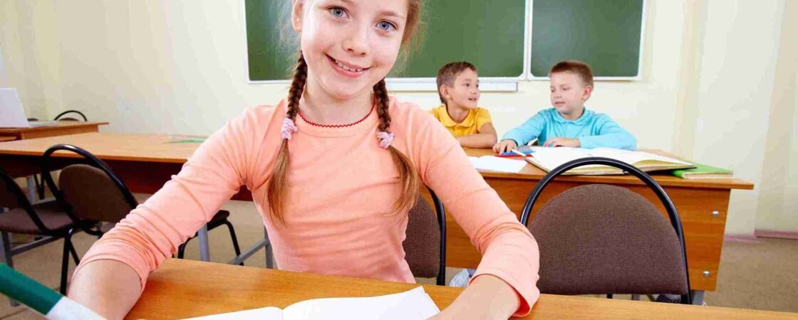 A young girl taking an 11 Plus Mock Exam at a desk in a classroom, with other students in the background.