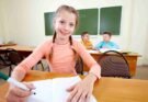 A young girl taking an 11 Plus Mock Exam at a desk in a classroom, with other students in the background.